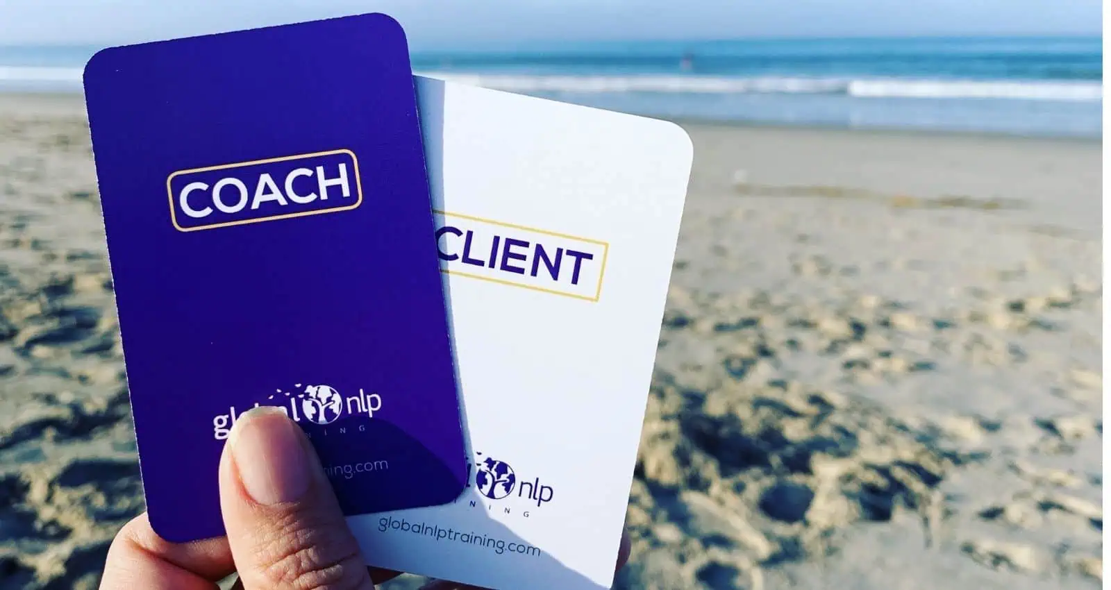 Hand holding Global NLP Training coach and client role-play cards on a beach